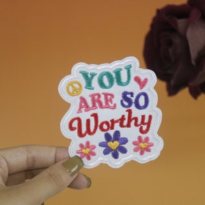Pode incluir: Etiqueta branca bordada com o texto "You are so worthy" em letras coloridas. A etiqueta é decorada com um sinal de paz, um coração e três flores.