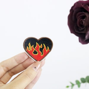 Pode incluir: Um remendo vermelho em forma de coração com chamas amarelas e laranja bordadas nele. O remendo é segurado em uma mão contra um fundo branco.