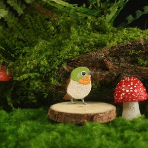 May include: A small, embroidered bird with green, white, and orange accents sits on a wooden log surrounded by three miniature mushrooms. Two of the mushrooms are red with white spots, and the third is black with white spots.