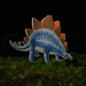 May include: Blue and brown embroidered iron-on patch of a dinosaur with a spiky back.