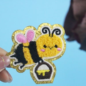 May include: A yellow and black embroidered bee with pink wings holding a brown honey pot. The bee has a cute cartoon face and is surrounded by a gold glitter border.