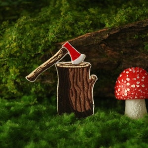 Könnte beinhalten: Ein roter und weißer Pilz mit weißen Punkten steht neben einem schwarzen und weißen Pilz mit weißen Punkten. Im Vordergrund befindet sich ein braunes gesticktes Stück einer Axt, die in einen Baumstumpf gesteckt ist.