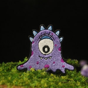 Peut inclure: Patch brodé violet d'un monstre borgne avec des taches roses et un sourire narquois.