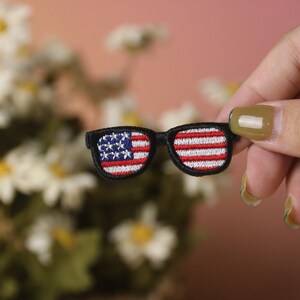 May include: A small embroidered patch of sunglasses with the American flag design. The sunglasses are black with red, white, and blue stitching.
