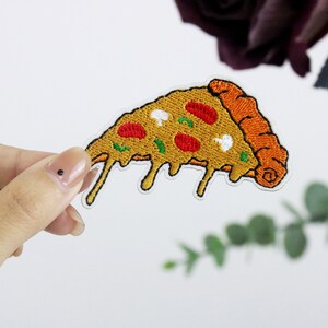 May include: A yellow and orange embroidered pizza slice with red pepperoni, green peppers, and white mushrooms. The pizza is dripping with melted cheese.