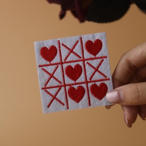 Puede incluir: Un tablero de fieltro blanco con un juego de tres en raya bordado en rojo. El tablero de juego tiene corazones rojos en lugar de las tradicionales X y O.