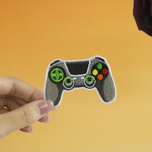 May include: A gray embroidered patch of a video game controller with green, yellow, red, and orange buttons.