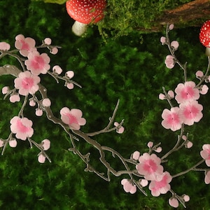 May include: Embroidered floral branch with pink blossoms and a silver-colored stem. The design is set against a backdrop of green moss, with decorative red and black spotted mushrooms.