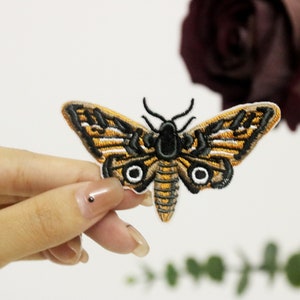 Può includere: Una toppa ricamata a forma di falena, di colore marrone e nero con accenti bianchi. La falena ha due grandi occhi bianchi.