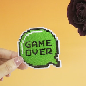 Op de afbeelding: Een groene, gepixelde spraakballon met de tekst "GAME OVER" in zwarte letters. De spraakballon wordt in een hand tegen een gele achtergrond gehouden.