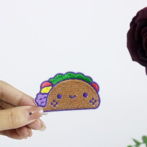 以下が含まれることがあります： 緑のレタス、赤と黄色の具材、紫の玉ねぎがついた、笑顔のタコの刺繍アイロン接着パッチです。