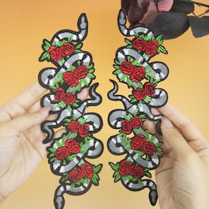 May include: Two embroidered iron-on patches featuring a snake intertwined with red roses. The snake is grey and white with black outlines. The roses are red with green leaves.