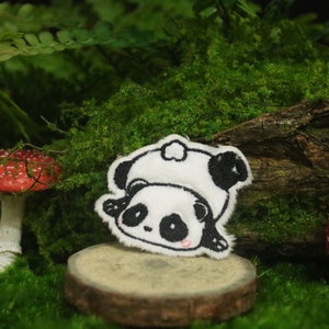 Puede incluir: Un parche bordado de panda blanco y negro con un sombrero blanco. El parche está sentado en un pequeño círculo de madera, rodeado de musgo verde.