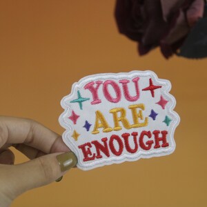 May include: White embroidered patch with the text "YOU ARE ENOUGH" in pink, red, yellow, and blue lettering. The patch is surrounded by colorful stars.