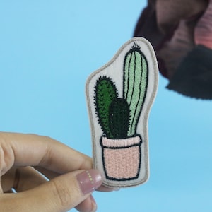 May include: A white felt patch with an embroidered cactus in a pink pot. The cactus is green and has two stems.