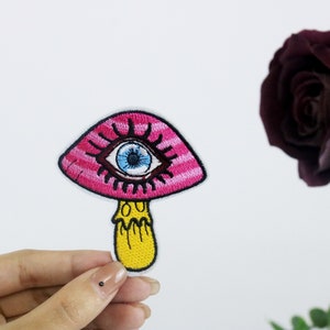 Könnte beinhalten: Ein rosa-weiß gestreifter Pilz mit einem blauen Auge und einem gelben Stiel. Der Pilz ist gestickt und wird von einer Hand gehalten.