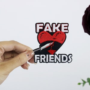 Op de afbeelding: Een geborduurde patch in zwart en rood met een hart dat door een mes is doorboord en de tekst "FAKE FRIENDS".