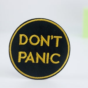 Könnte beinhalten: Ein runder, schwarzer gestickter Aufnäher mit goldener Umrandung und dem Text "DON'T PANIC" in Gold.