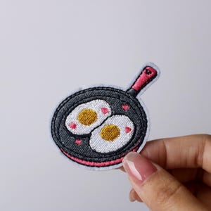 May include: An embroidered iron-on patch depicting a frying pan with two sunny-side-up eggs. The pan is black and white with a red handle and pink hearts around the eggs.