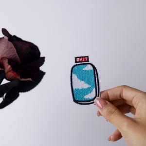 May include: A blue and white embroidered patch with a cloud design and the word "EXIT" in red. The patch is held in a hand.