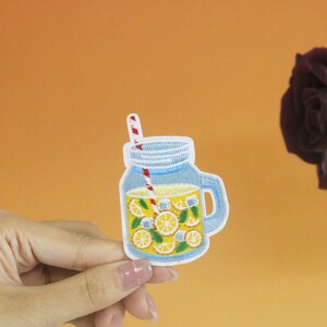 Peut inclure: Un bocal en verre bleu avec une paille rouge et blanche rayée rempli de limonade jaune et de tranches de citron. Le bocal a un couvercle blanc et une poignée.