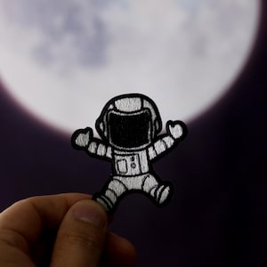 Puede incluir: Un astronauta blanco con contornos negros y una visera negra está sosteniendo sus brazos frente a él. El astronauta está sobre un fondo oscuro con una gran luna blanca y brillante en el fondo.