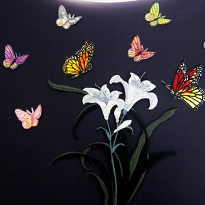 Könnte beinhalten: Ein schwarzer Stoffhintergrund mit weißen Lilien und bunten Schmetterlingen. Die Schmetterlinge sind in verschiedenen Farben, darunter Gelb, Orange, Rosa, Blau und Rot.