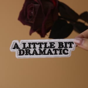 May include: White iron-on patch with black embroidered text that reads "A little bit dramatic".