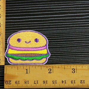 May include: A yellow, purple, pink, and green embroidered iron-on patch of a smiling cartoon hamburger. The patch is about 1.5 inches wide.