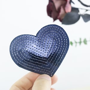 Può includere: Una toppa a forma di cuore blu scuro realizzata con paillettes. Le paillettes sono disposte in un motivo circolare, creando un effetto vorticoso.