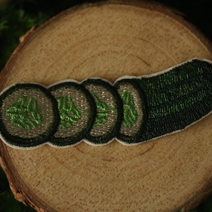 May include: Embroidered patch depicting a sliced cucumber. The patch shows a dark green cucumber with four circular slices, each with a green embroidered design. The patch is set against a wooden background.
