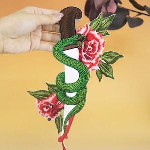 May include: Green snake wrapped around a white dagger with a brown handle. Red roses with green leaves are on either side of the dagger. The background is a light orange color.