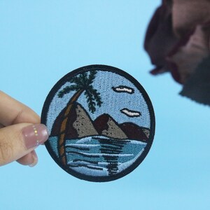 Puede incluir: Un parche redondo bordado con un fondo azul que representa una palmera en una pequeña isla con montañas marrones y agua azul. El cielo es azul claro con nubes blancas.