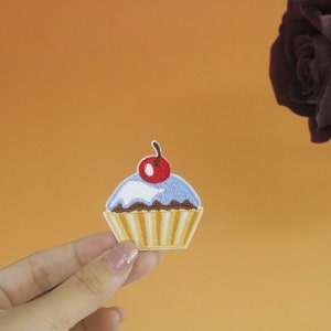 May include: A blue and yellow embroidered cupcake patch with a red cherry on top. The patch is held in a hand against a light orange background.