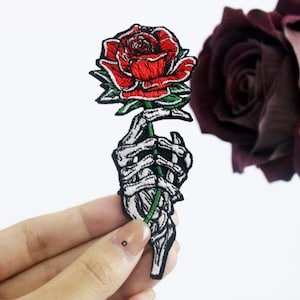 May include: An embroidered iron-on patch featuring a skeleton hand holding a red rose. The rose is detailed with green leaves and a black stem.