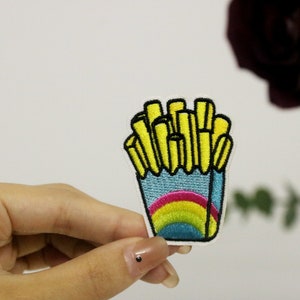以下が含まれることがあります： 青い容器に入ったフライドポテトの漫画の刺繍パッチ。虹のデザイン。フライドポテトは黄色で黒いアウトライン。パッチは手で持たれています。