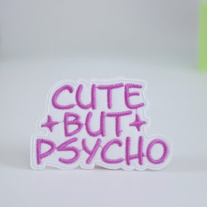 May include: A white embroidered patch with the phrase "CUTE BUT PSYCHO" in pink lettering. The patch has a white border and star-like accents. The patch is on a white surface.