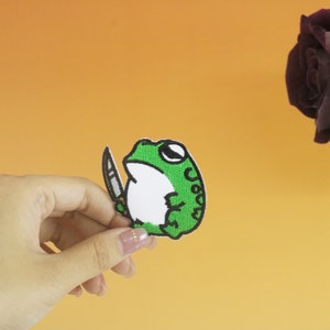 以下が含まれることがあります： 手にナイフを持った白いお腹の緑色のカエル。カエルはパッチに刺繍されています。