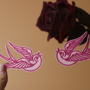 Puede incluir: Dos parches bordados de color rosa que representan golondrinas volando. Los parches están sobre un fondo marrón claro.