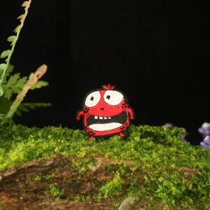 以下が含まれることがあります： 白い目で大きく開いた口、黒い輪郭の漫画のモンスターの赤い刺繍パッチ。モンスターは緑の苔むした表面にあります。