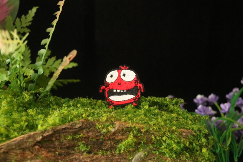 以下が含まれることがあります： 白い目で大きく開いた口、黒い輪郭の漫画のモンスターの赤い刺繍パッチ。モンスターは緑の苔むした表面にあります。