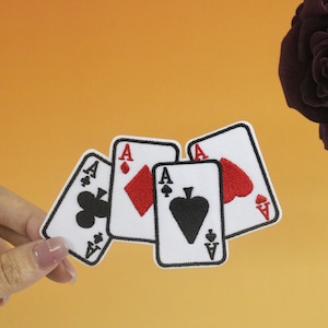 May include: Four embroidered patches of playing cards, each featuring the Ace of a different suit: clubs, diamonds, spades, and hearts. The patches are white with black and red embroidery.