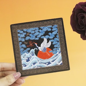 Op de afbeelding: Geborduurde patch met een witte kraan die over een rode zon en blauwe golven vliegt. De patch is omgeven door een zwart en gouden frame met een Grieks sleutelpatroon.