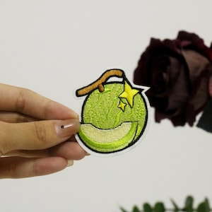 Op de afbeelding: Een groene meloen met een bruine steel en twee gele sterren geborduurd op een witte achtergrond. De meloen is een populair fruit in Japan en wordt vaak gebruikt in desserts en dranken.