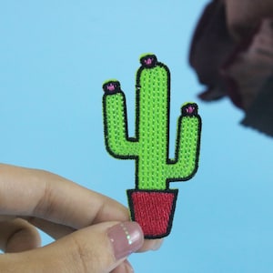 May include: A green cactus with pink flowers in a red pot. The cactus is embroidered on a fabric patch.