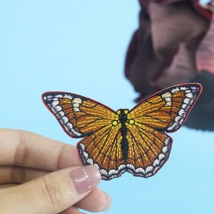 May include: A brown and yellow embroidered butterfly patch with white and purple accents. The patch has a red outline and is held in a hand.
