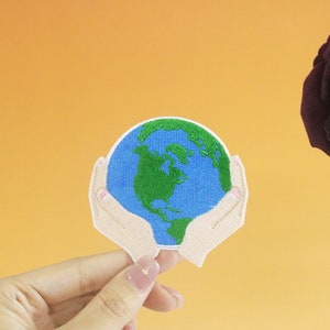 May include: A hand-stitched embroidered patch depicting a pair of hands holding a blue and green globe. The globe is a representation of the Earth with continents outlined in green.