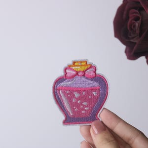 May include: A pink and purple embroidered iron-on patch of a heart-shaped bottle with a pink bow and a yellow lid. The bottle is filled with pink glitter.