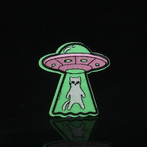 Puede incluir: Un parche bordado que representa un gato siendo abducido por un OVNI, en verde y rosa.