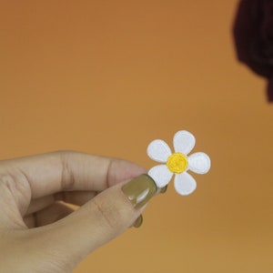 以下が含まれることがあります： 白い花びらと黄色の芯を持つ、刺繍された白いデイジーの花。花は手で持たれている。